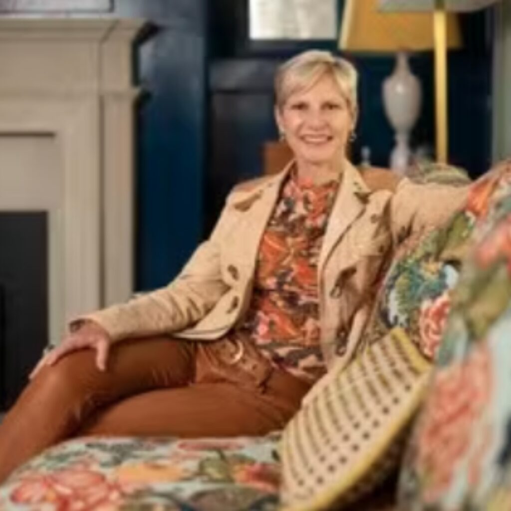 A woman with short blonde hair, wearing a floral top and beige jacket, sits on a patterned sofa in a well-decorated living room.