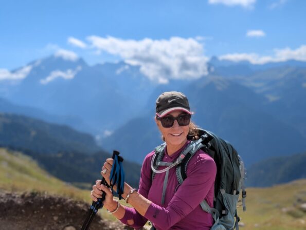 A woman wearing sunglasses and a backpack stands on a mountain trail, holding trekking poles, with mountains and a blue sky in the background.
