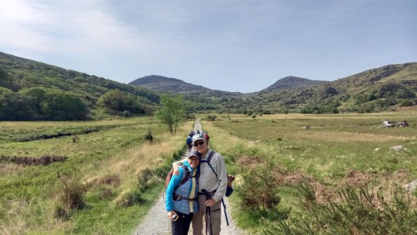 Two people wearing hiking gear stand together on a trail surrounded by grassy hills and mountains under a clear sky.