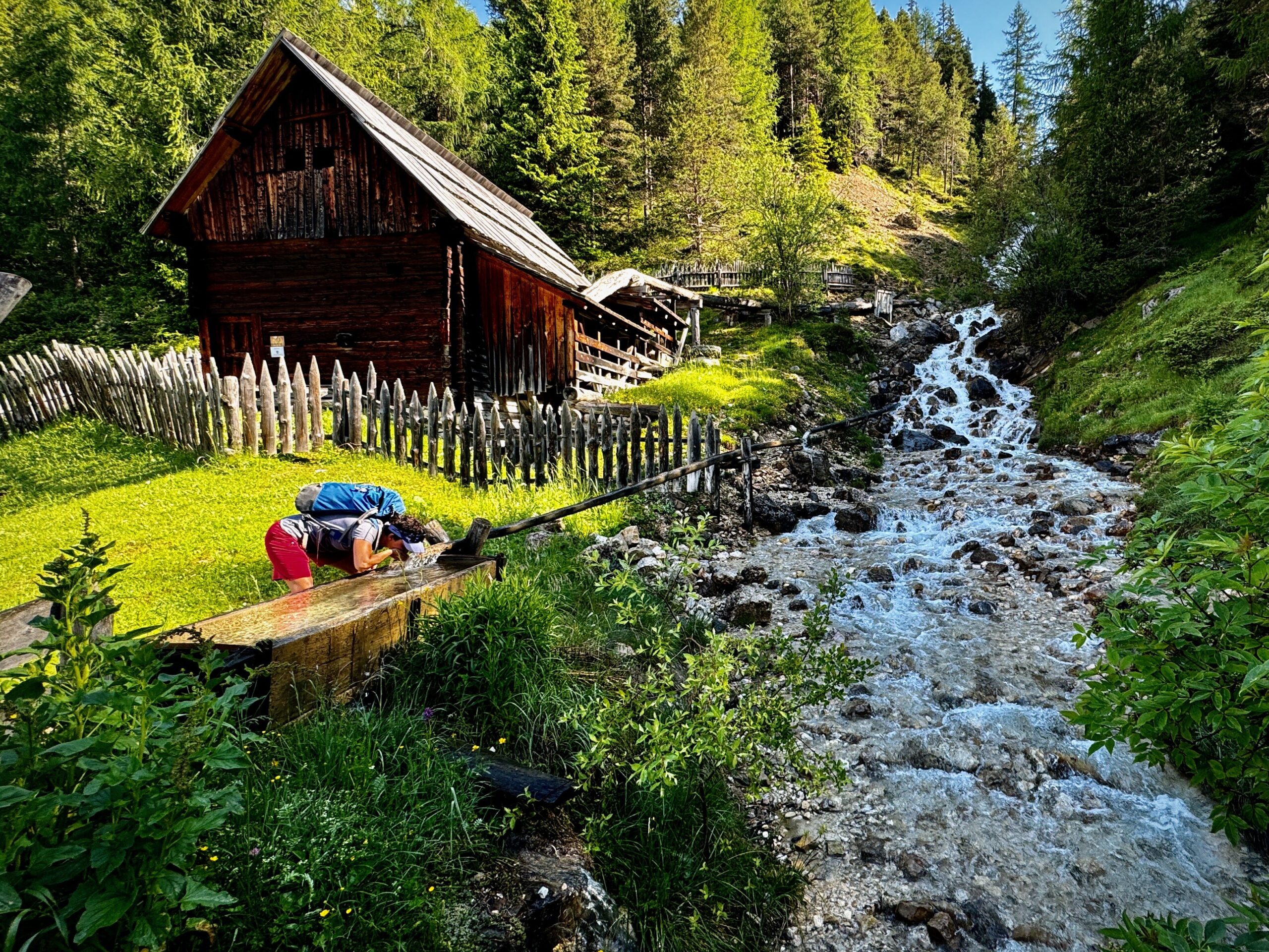 A person bends over a wooden trough near a flowing stream, beside a rustic wooden cabin and fence in a lush, green forested area.