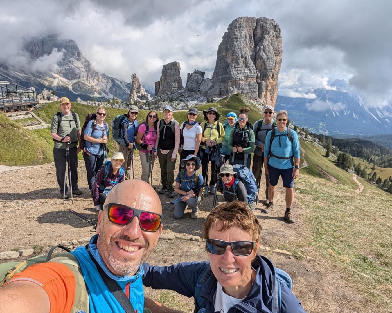 A group of hikers poses for a photo on a mountain trail with rocky peaks and cloudy skies in the background.