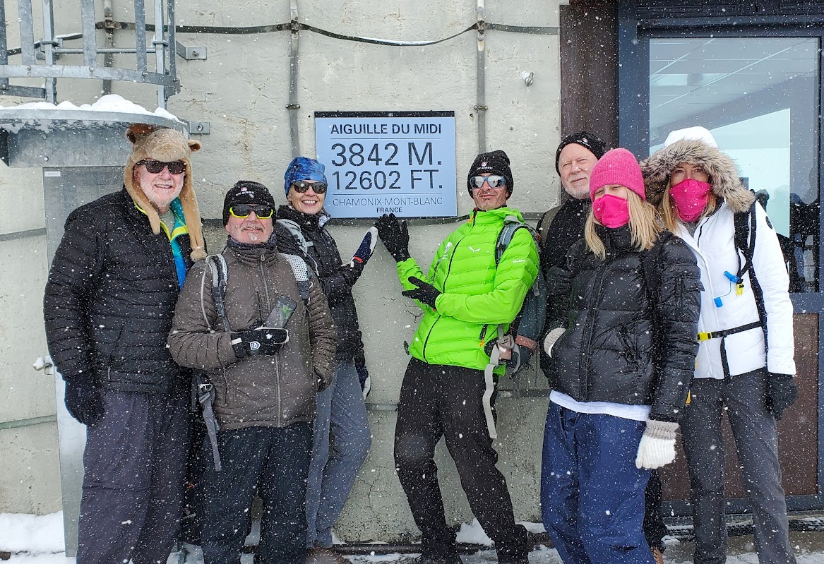 A group of seven people in winter clothing stand in the snow beside a sign reading "Aiguille du Midi 3842 M 12602 FT"—a perfect moment from their Alps hiking adventure.