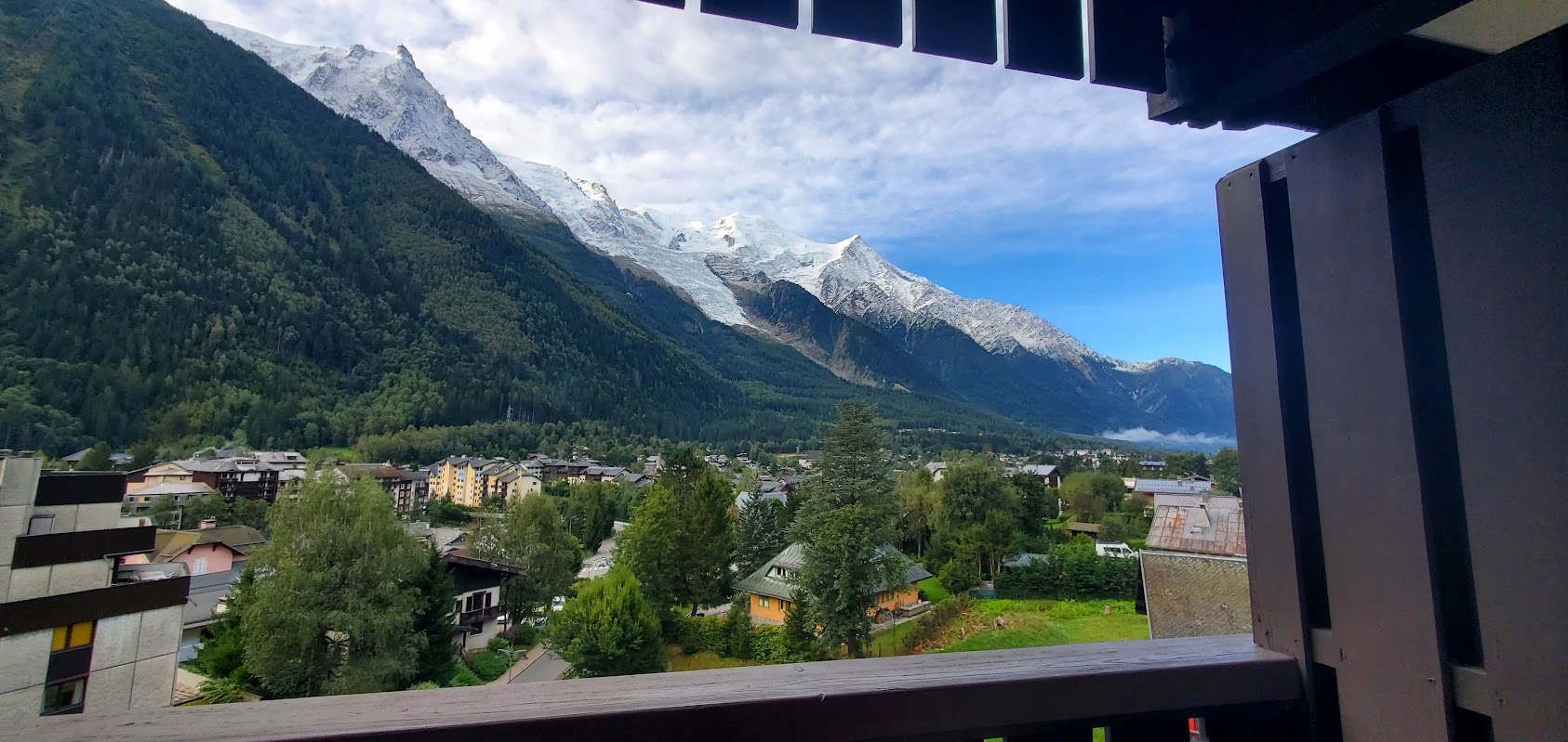 View from a balcony overlooking a town with trees and buildings, set against forested hills and snow-capped Alps—perfect for alp hiking or planning your next Chamonix and Zermatt hiking adventure under a partly cloudy sky.