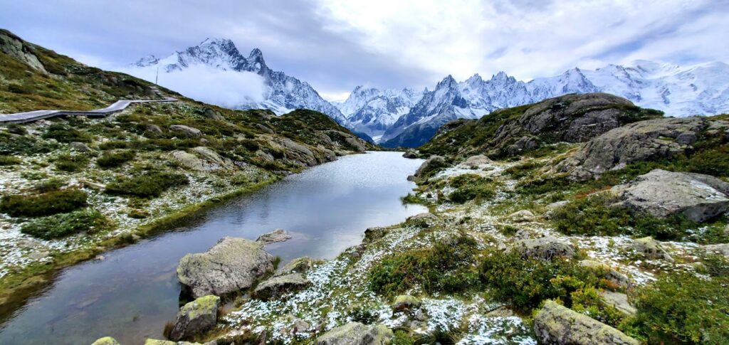 Alpine landscape featuring a clear pond surrounded by rocky terrain, green vegetation, and distant snow-capped mountains under a cloudy sky—perfect for alps hiking or exploring popular routes like Chamonix and Zermatt hiking.