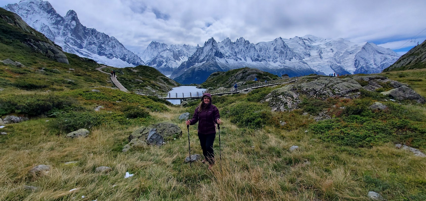 A person with hiking poles stands on a grassy trail, enjoying classic Alps hiking with a lake, footbridge, and snow-capped mountains in the background under a cloudy sky.