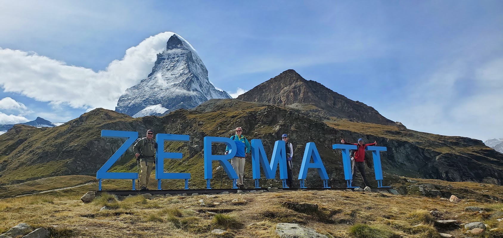 Four people stand by large blue “ZERMATT” letters on a grassy hill, with the Matterhorn towering in the background—a perfect snapshot of Chamonix and Zermatt hiking adventures in the Alps under a partly cloudy sky.