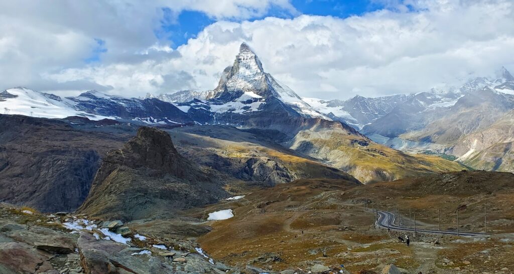 A view of the Matterhorn mountain with snow on its peak, surrounded by rocky terrain and patches of snow under a partly cloudy sky, perfect for alp hiking adventures.