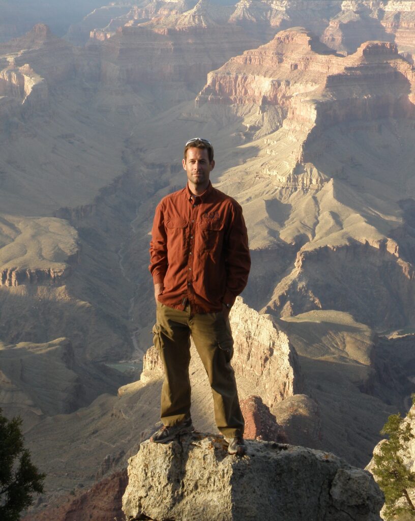 A person in a brown jacket and khaki pants stands on a rock ledge overlooking the vast landscape of the Grand Canyon, embodying the spirit of adventure featured in recent Mt Sobek news.