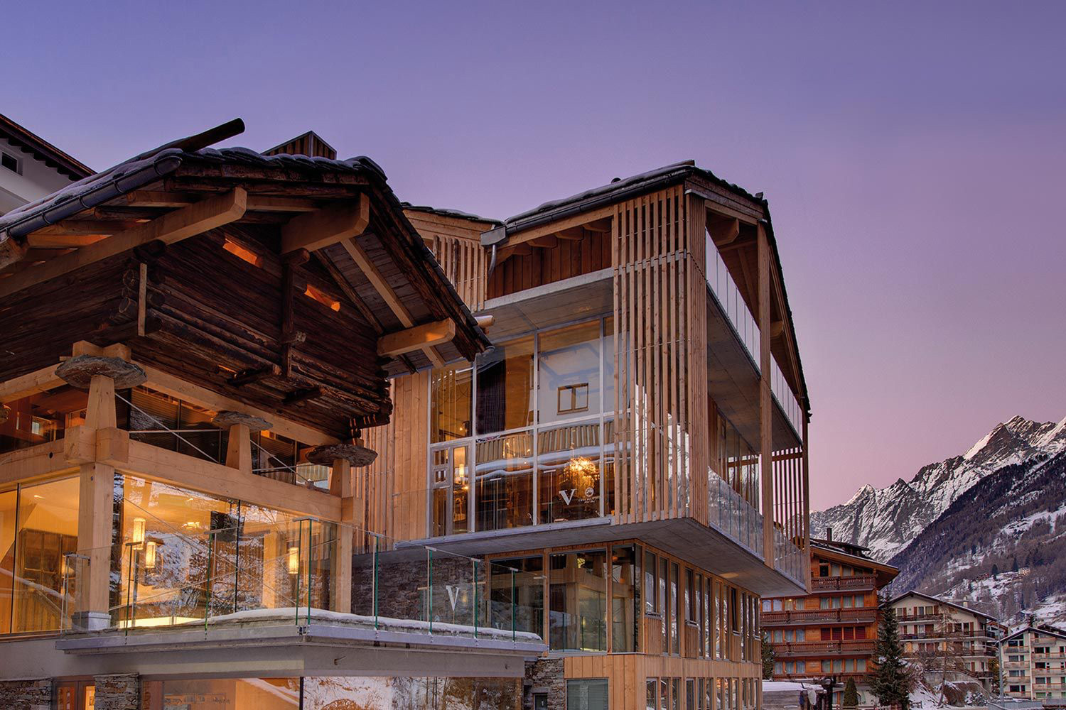 A modern wooden and glass building stands next to a traditional chalet, with snow-capped mountains and other buildings visible in the background at dusk—a perfect scene for alps hiking enthusiasts.