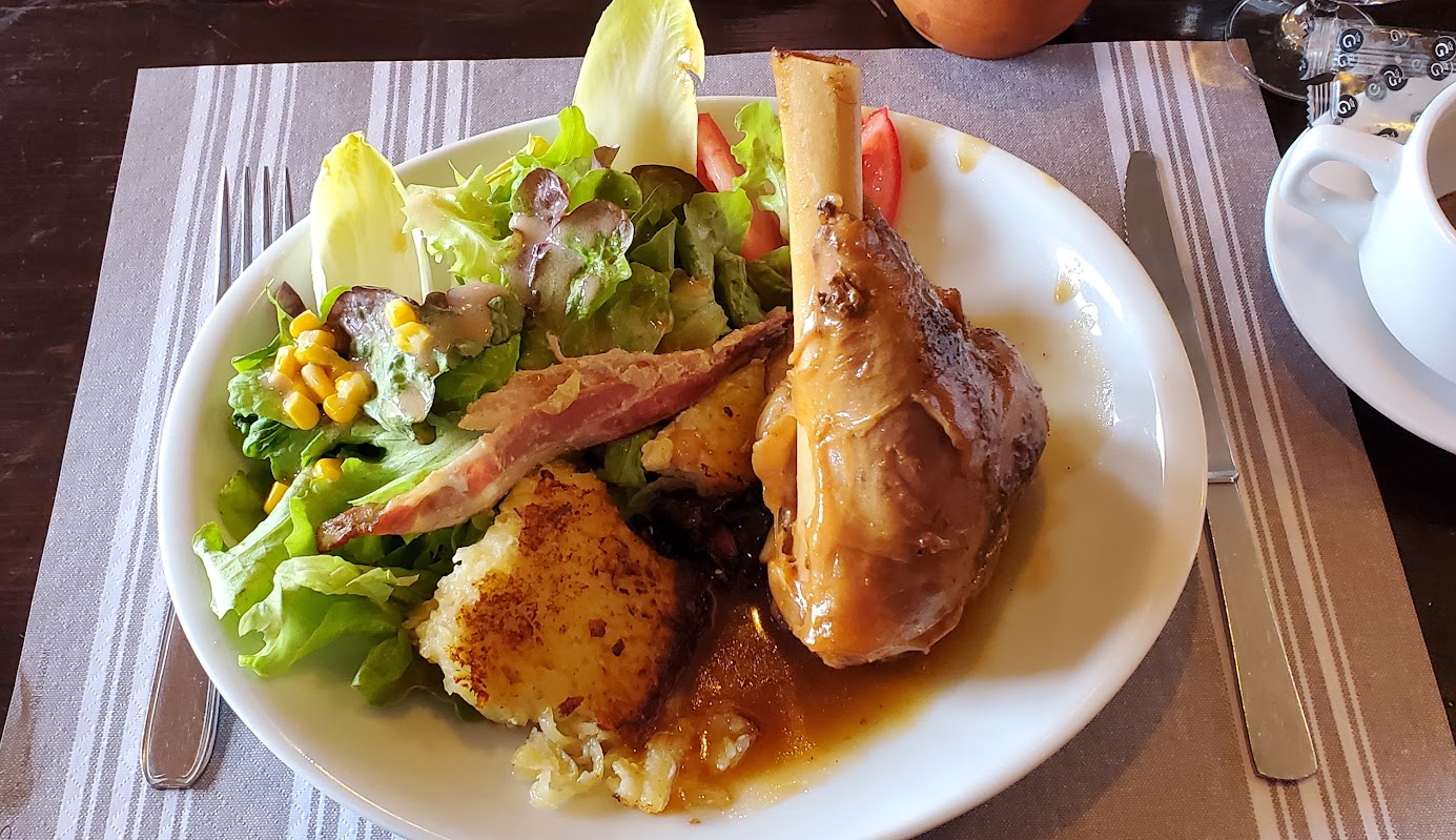 A plate with braised meat on the bone, leafy green salad with corn and dressing, a slice of bacon, and cheesy potatoes—perfect for refueling after a day of alps hiking or exploring Chamonix and Zermatt hiking trails.