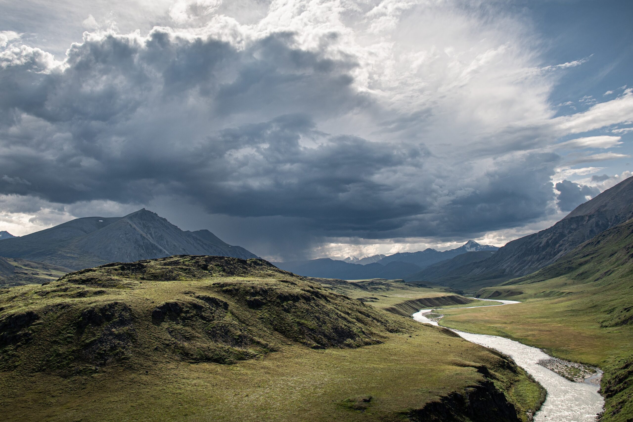 A winding river flows through a green valley with hills and mountains under a dramatic sky filled with dark storm clouds, creating a scene perfect for adventurous Alaska tours.
