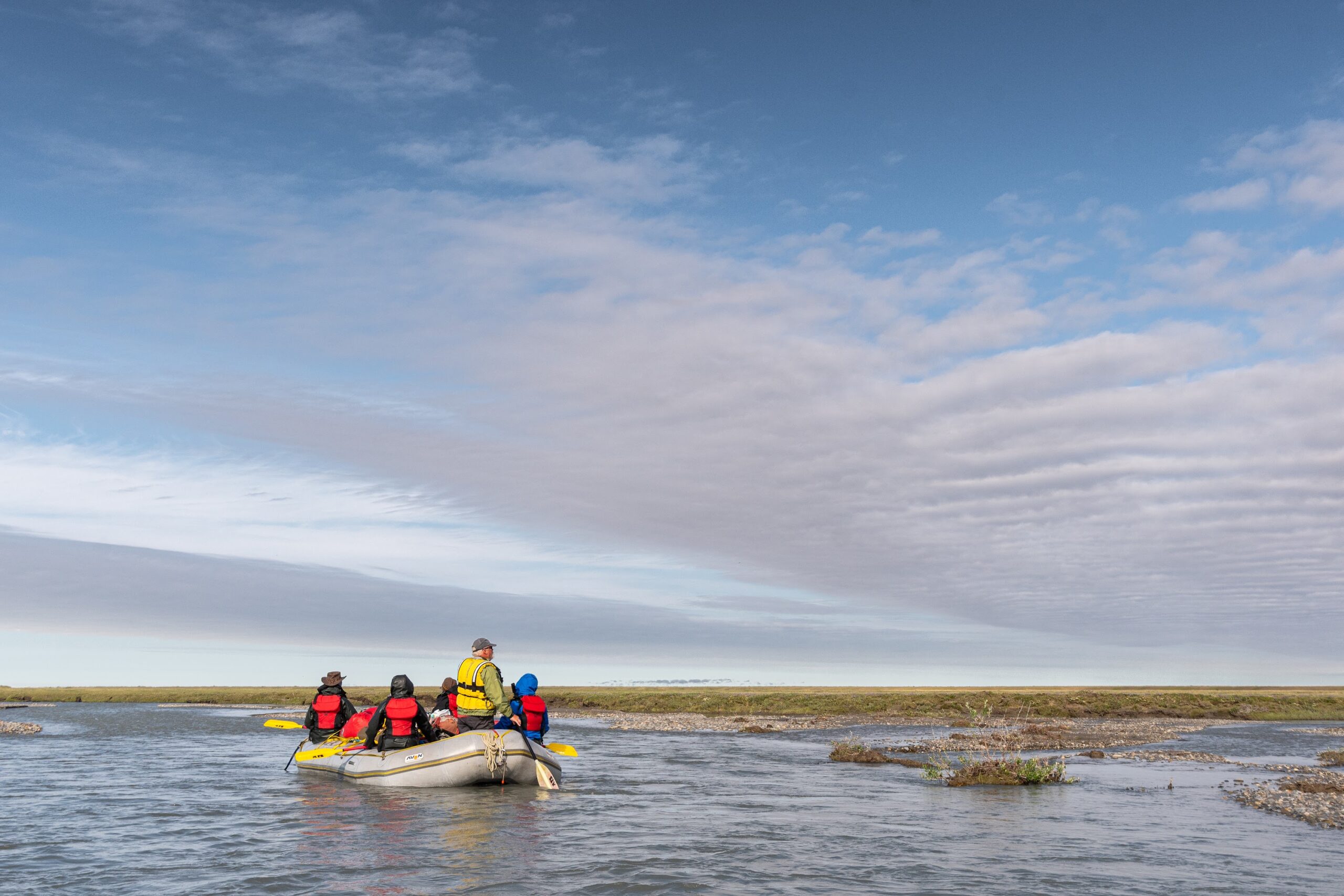 A group of people wearing life jackets sit in an inflatable raft on a calm river, enjoying one of the many scenic Alaska tours under a partly cloudy sky with open grassy land in the background.