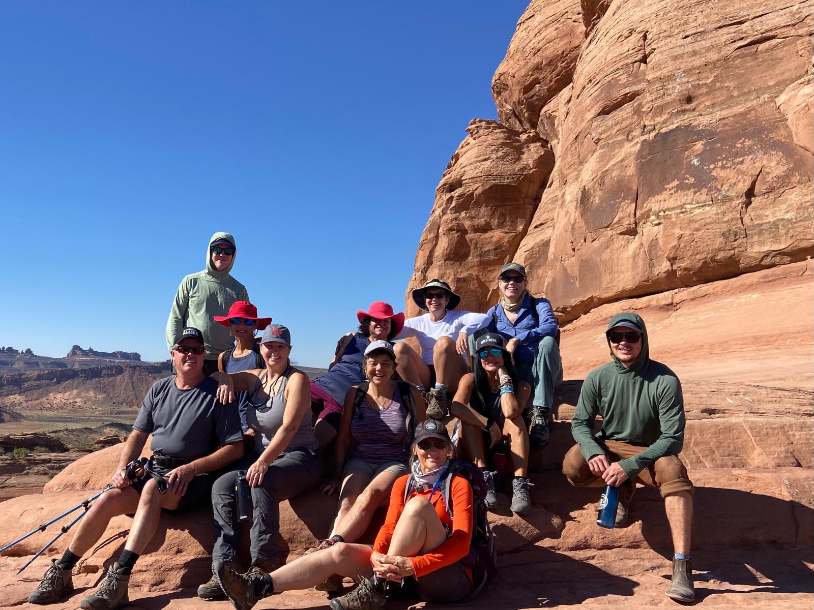 A group of people in hiking attire sit and pose together on red rock formations under a clear blue sky, capturing the adventurous spirit of Utah national parks hiking.