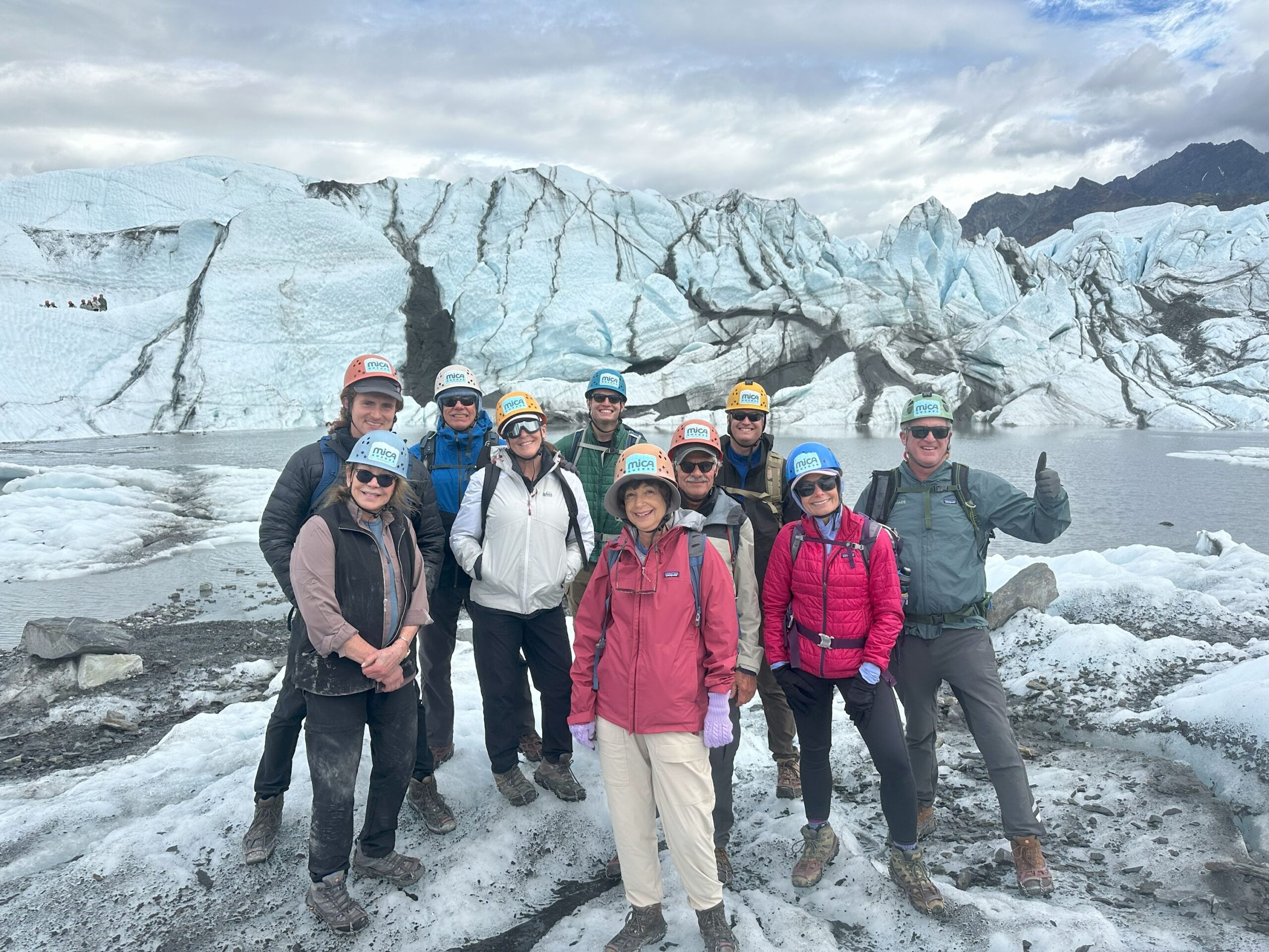 A group of people wearing helmets and outdoor gear stand on a glacier with icy terrain and mountains in the background, ready to embark on one of the thrilling Alaska tours.