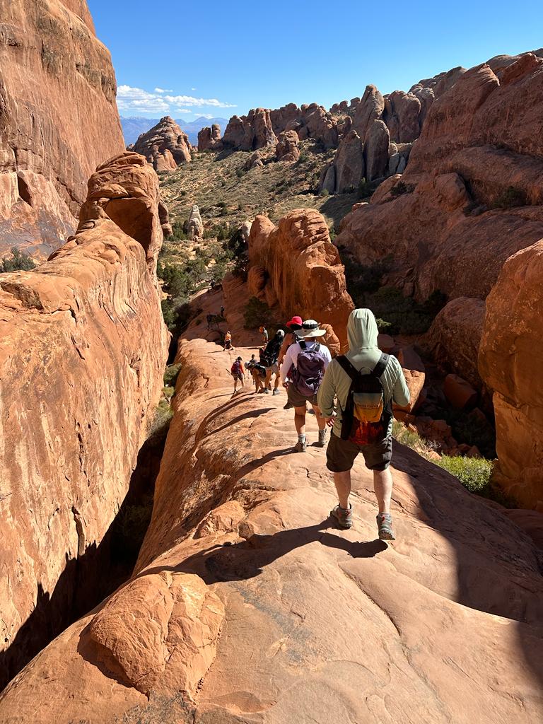 A group of hikers walk along a narrow rocky trail surrounded by large red rock formations under a clear blue sky, capturing the classic beauty of Utah national parks hiking.