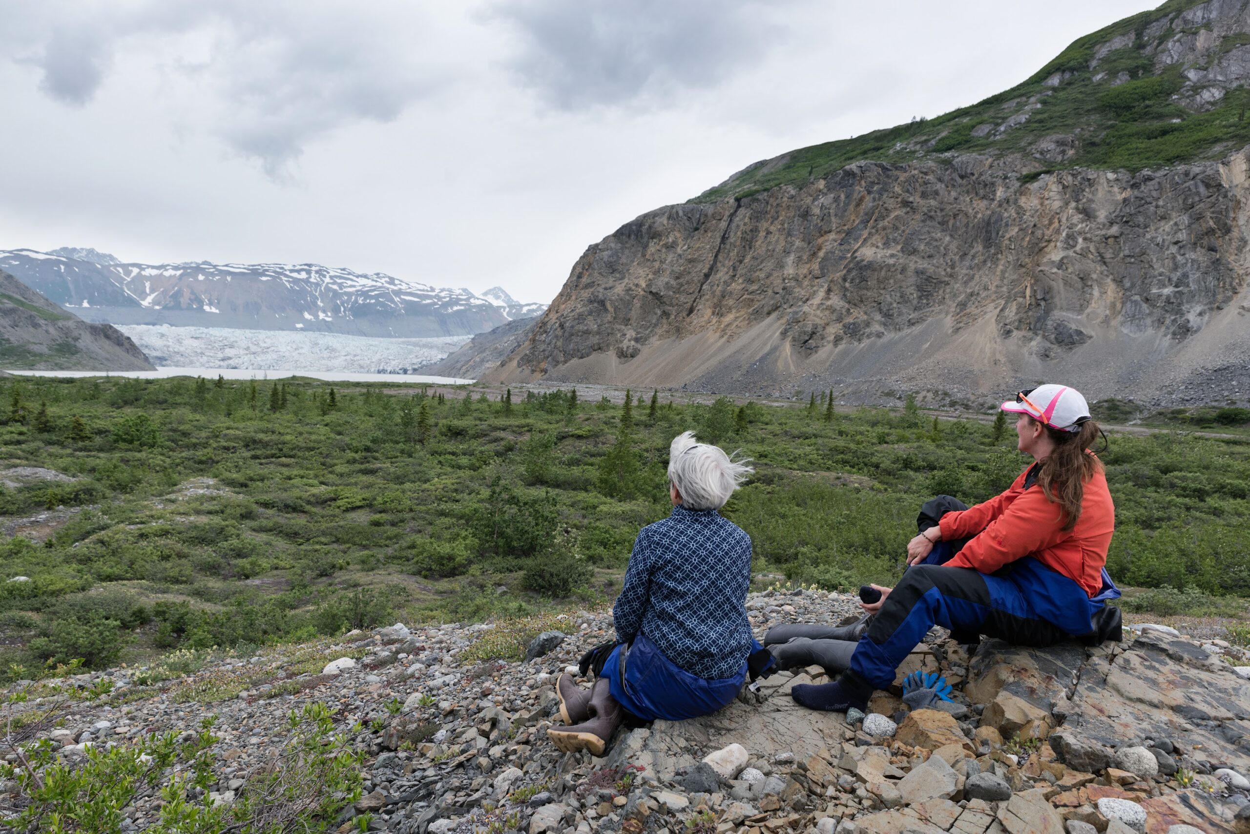 Two people sit on rocky ground overlooking a green valley with a glacier and snow-capped mountains in the distance under a cloudy sky, capturing the breathtaking beauty often seen on Alaska tours.
