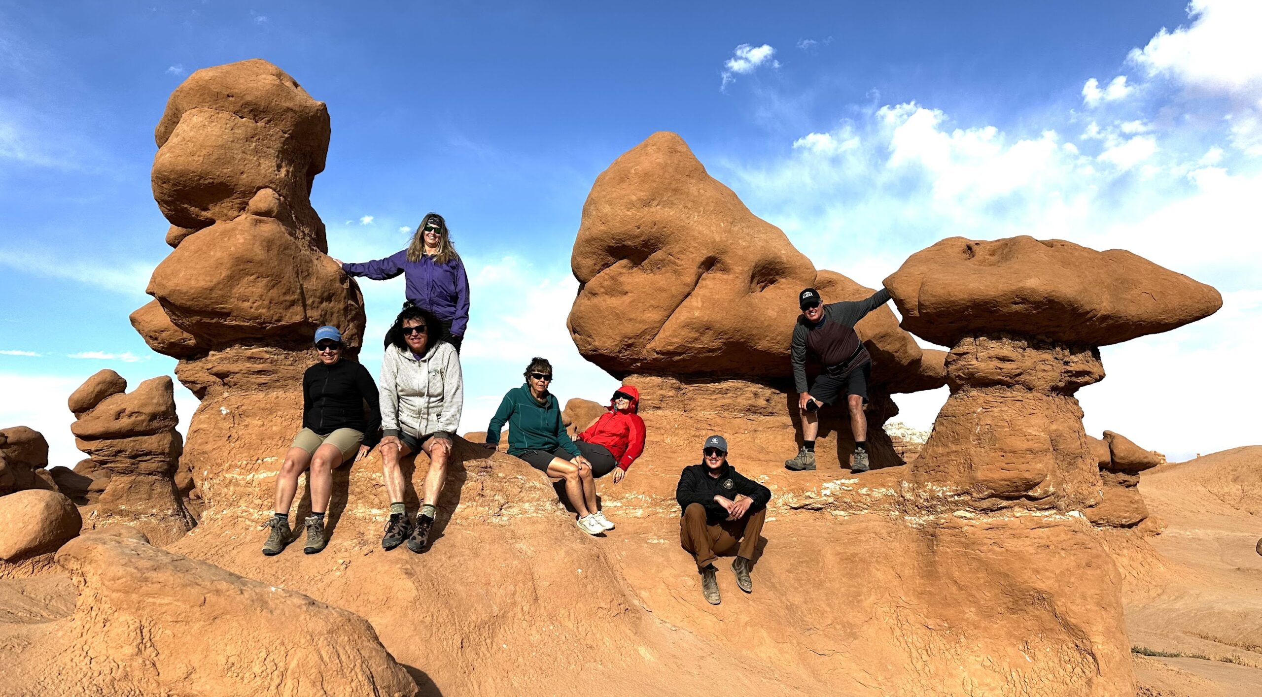 Six people sit and pose on large, uniquely shaped red rock formations under a blue sky with scattered clouds, capturing the adventurous spirit of hiking in Utah national parks.