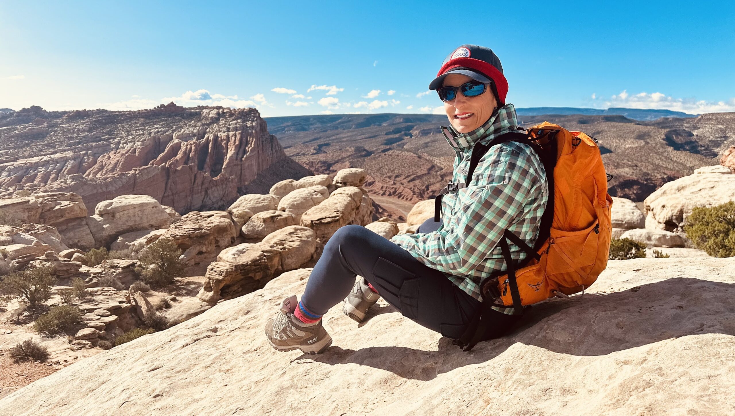 Person wearing outdoor gear and an orange backpack sits on a rocky ledge overlooking a desert canyon landscape in Utah national parks under a clear blue sky, capturing the spirit of adventure and hiking.