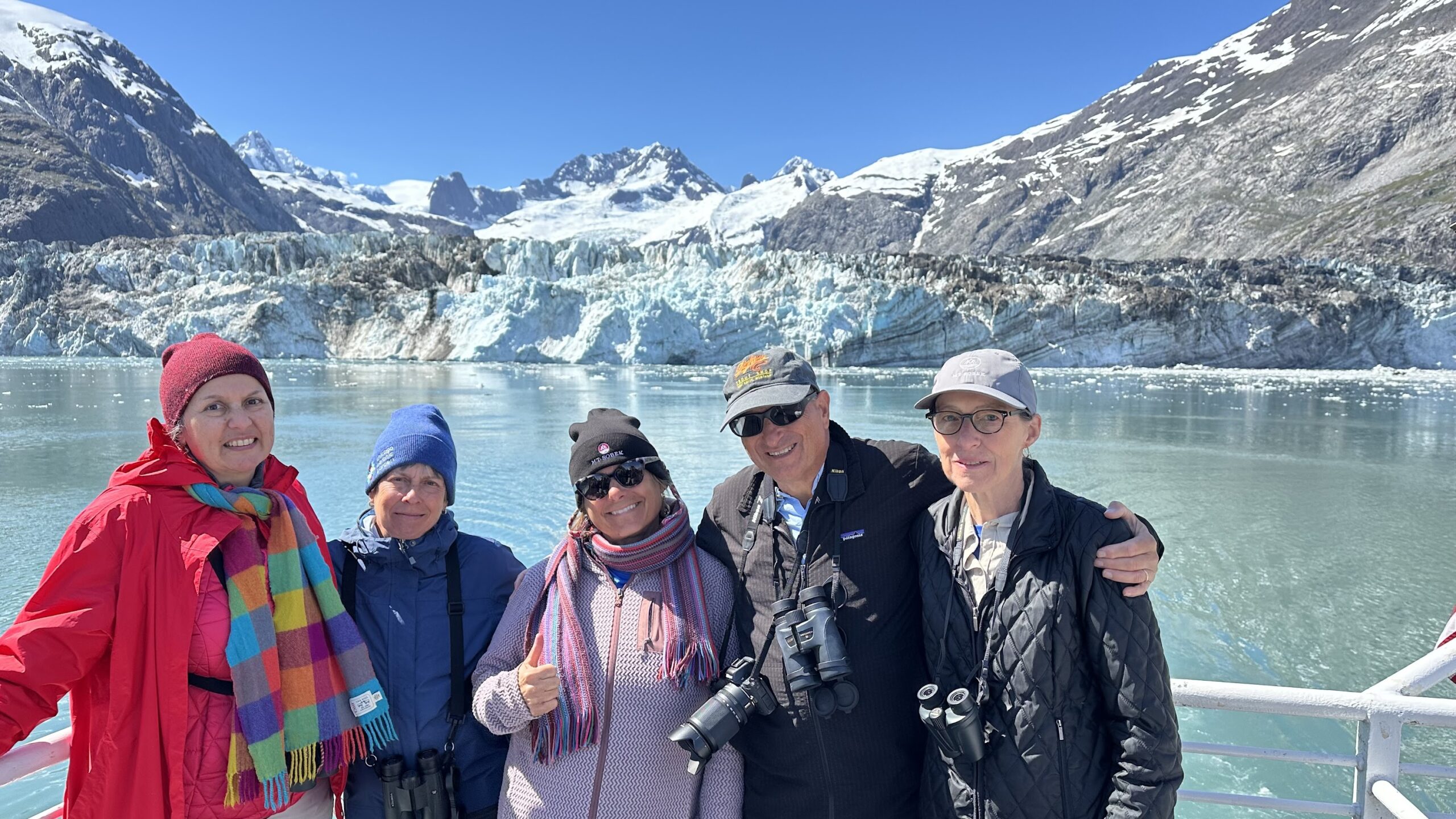 Five people in outdoor clothing stand on a boat during one of their Alaska tours, with snowy mountains and a glacier in the background, smiling at the camera.