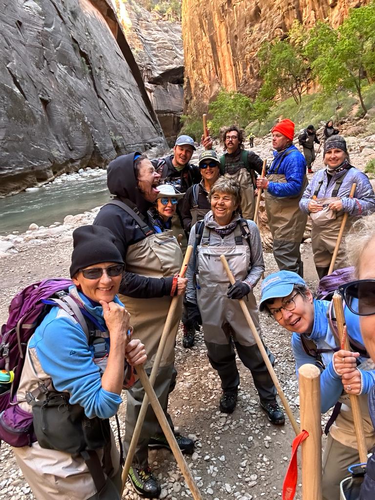 A group of people in waders and outdoor gear stand on a rocky riverside trail, smiling at the camera during their Utah national parks hiking adventure. Towering canyon walls and lush green trees rise behind them.
