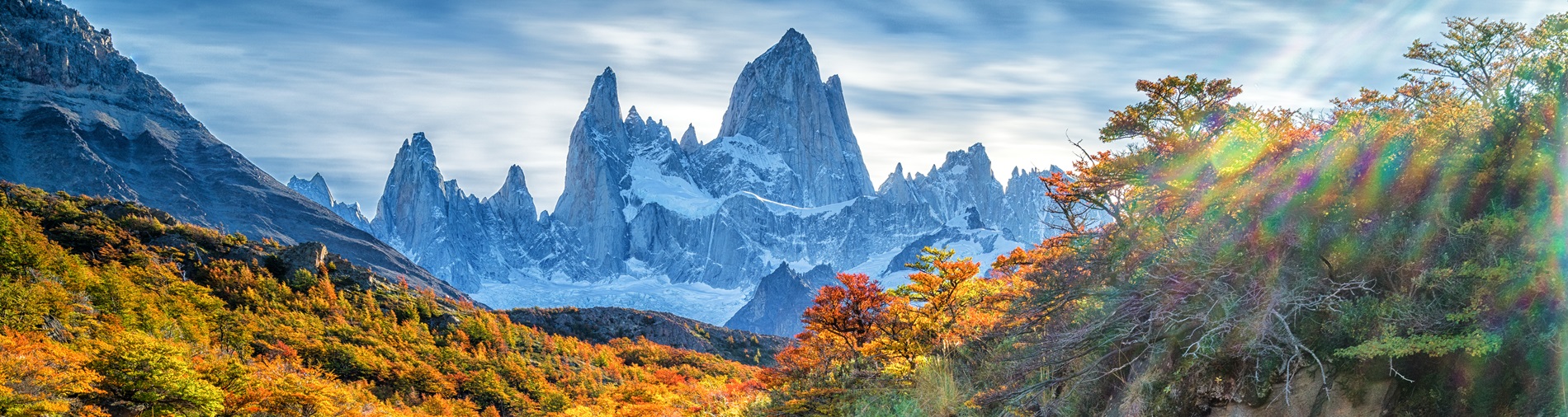 Jagged mountain peaks rise above a vibrant autumn forest, with sunlight streaming through trees and illuminating the foliage.