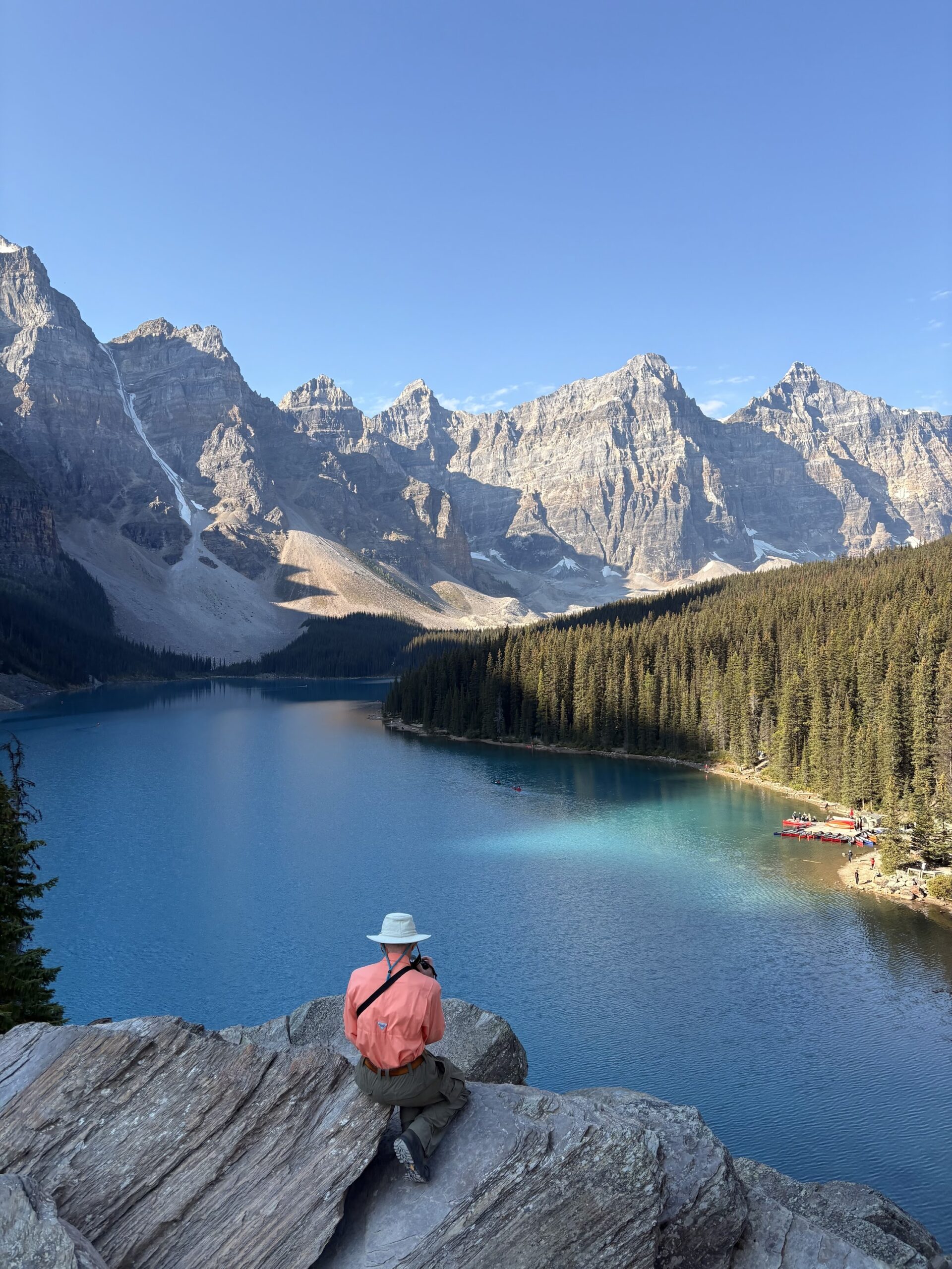 Person in a hat and orange jacket sits on a rock overlooking a blue lake surrounded by pine trees and the tall, rocky mountains that make the best MT Sobek Canadian Rockies hiking tours unforgettable under a clear blue sky.