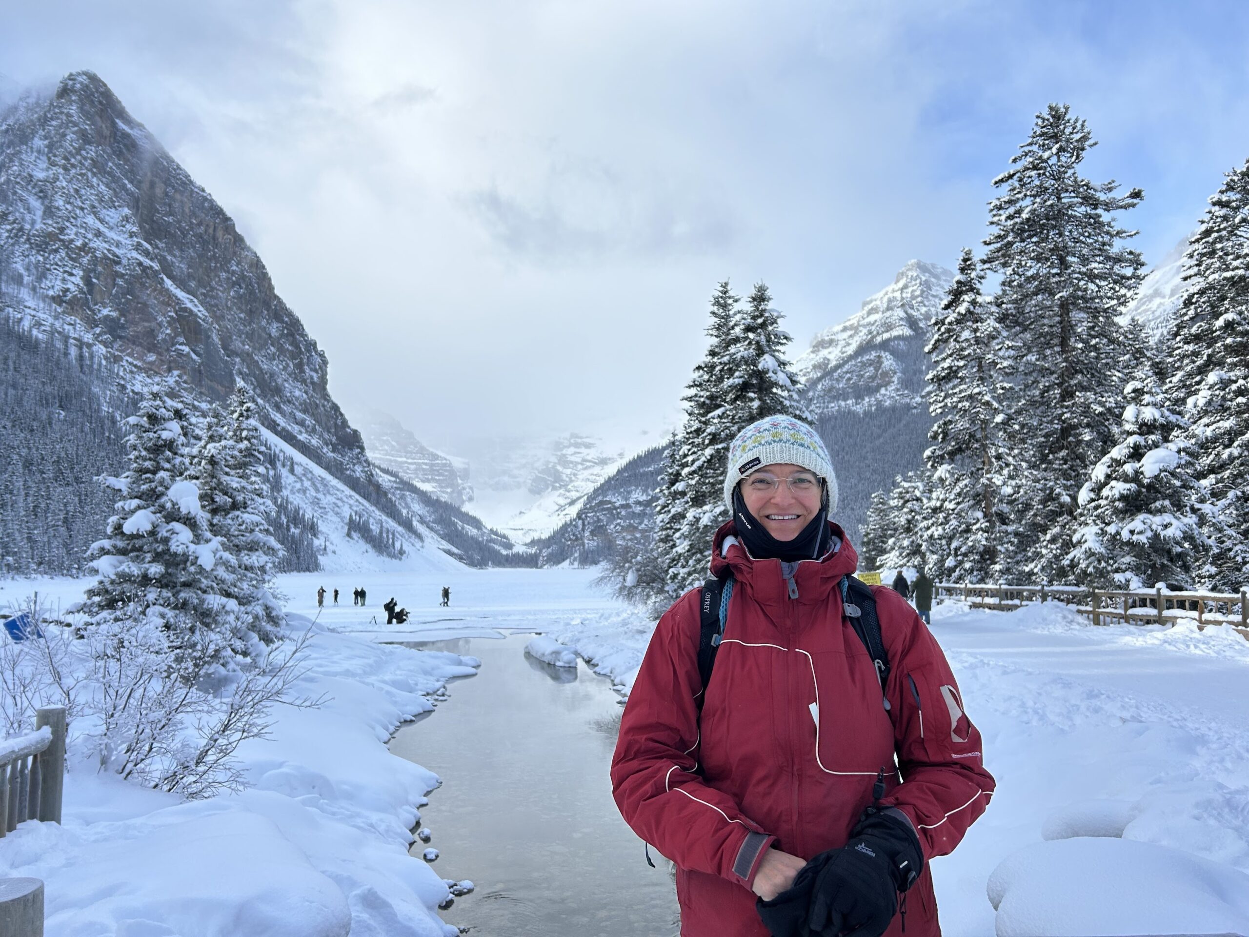 A person in a red winter jacket and white knit hat stands in front of a snowy mountain landscape, a partially frozen river, and pine trees—capturing the beauty found on the best MT Sobek Canadian Rockies hiking tours.