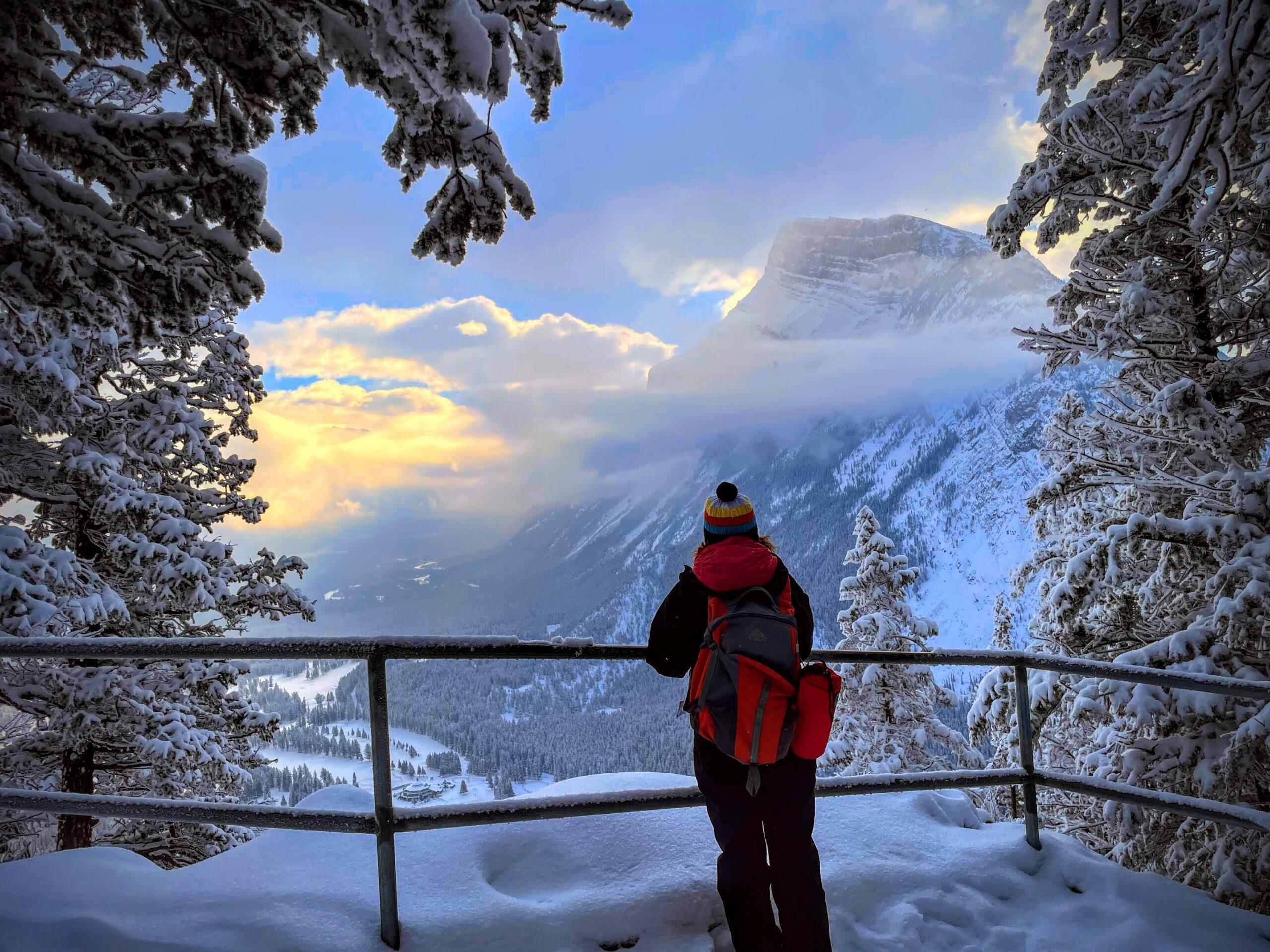 Bundled in winter clothing, a person stands at a snowy overlook, gazing at a mountain landscape lit by sunset—an experience reminiscent of the best MT Sobek Canadian Rockies hiking tours.