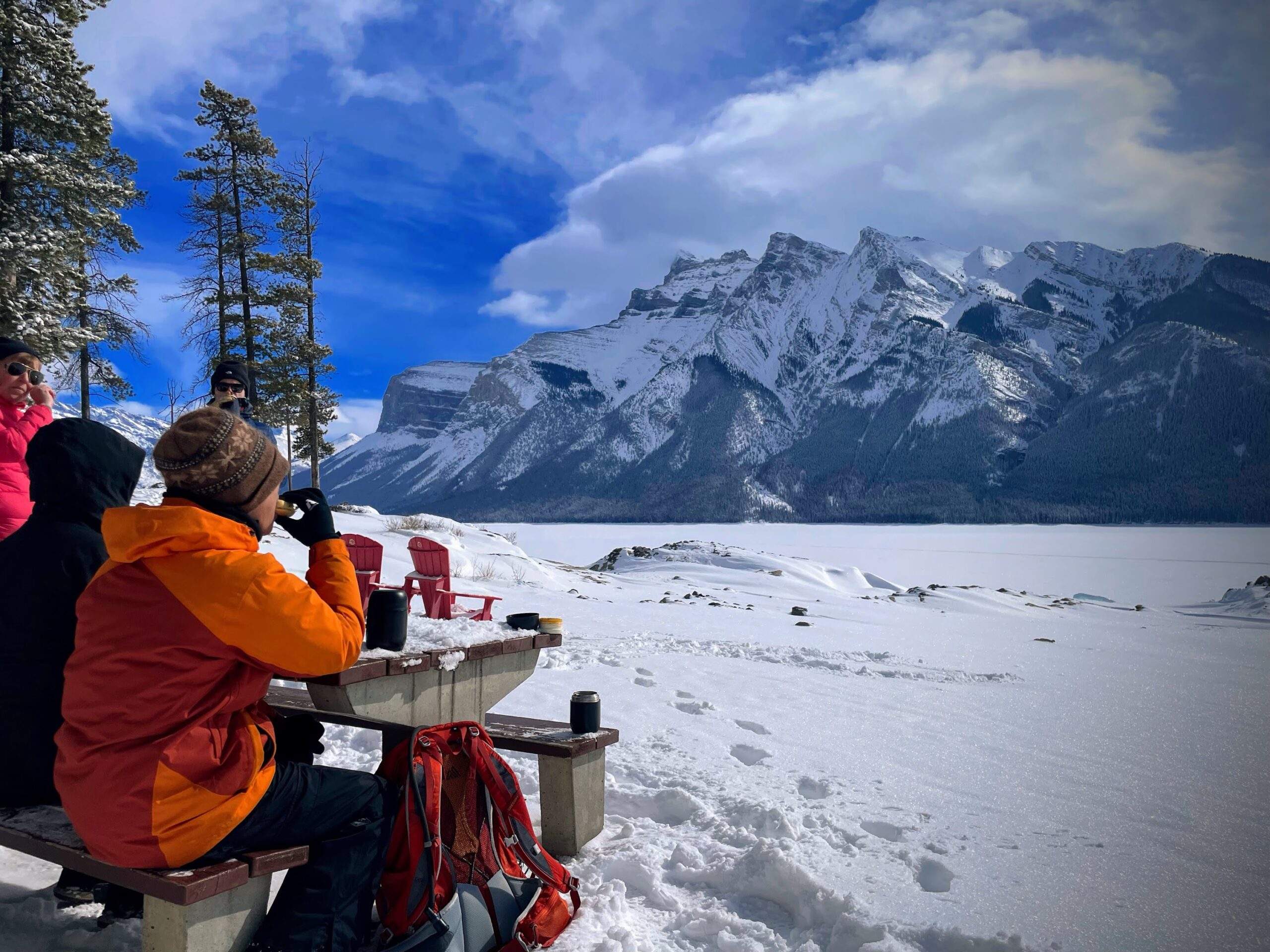 People in winter jackets sit at a picnic table on snow, drinking and talking, with snowy mountains and a frozen lake in the background—an ideal stop during one of the best MT Sobek Canadian Rockies hiking tours.